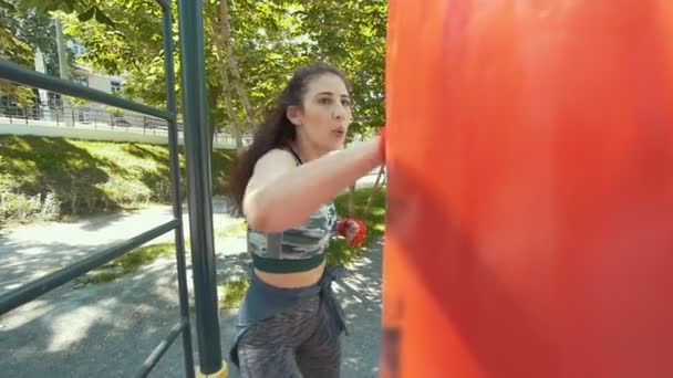 Mujer joven deportiva en ropa deportiva supera a un saco de boxeo, entrenamiento en el parque de verano, cámara lenta — Vídeos de Stock