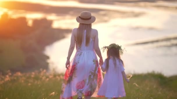 Visão traseira da jovem mãe de chapéu e pequena filha de mãos dadas desce a colina no pôr do sol de verão — Vídeo de Stock