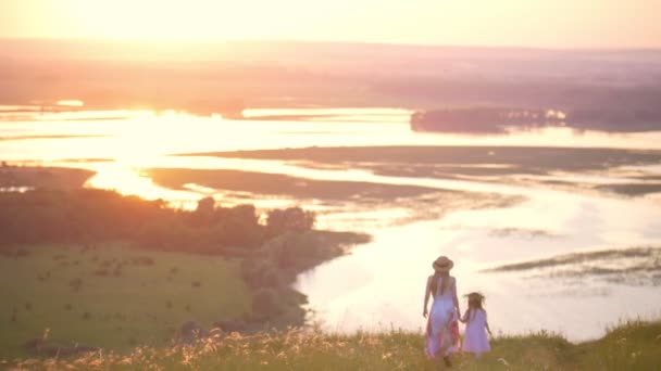 Visão traseira da mãe e da filhinha de mãos dadas desce a colina com vista perfeita sobre o rio ao pôr do sol — Vídeo de Stock