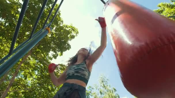 Mujer joven atlética en ropa deportiva supera a un saco de boxeo, entrenamiento en el parque de verano - vista inferior, cámara lenta — Vídeo de stock