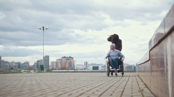 Homem deficiente em uma cadeira de rodas com jovem caminhando na rua da cidade — Vídeo de Stock