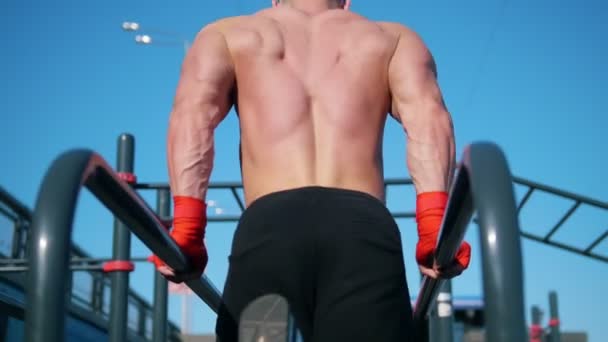 Young muscular sportsman pushing up at the parallel bars - workout at summer outdoor - rear view — Stock Video