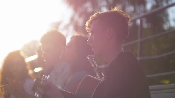 Jóvenes amigos cantando canciones con una guitarra en el parque al atardecer — Vídeos de Stock