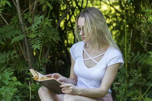 Jovem mulher lendo um livro sentado nos arbustos no dia de verão — Fotografia de Stock