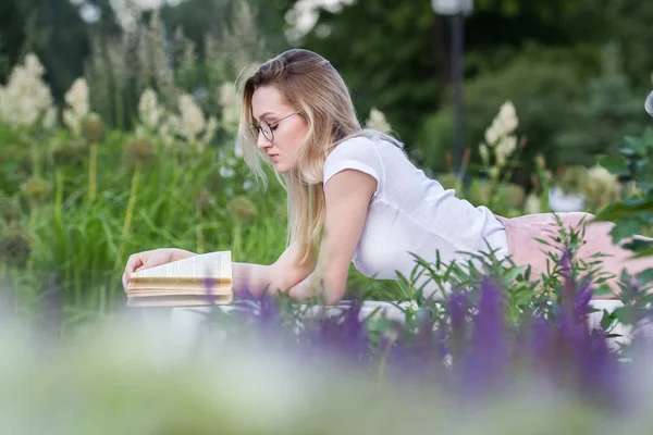 Jovem mulher bonita lendo um livro deitado no banco em torno da grama e flores no dia de verão — Fotografia de Stock