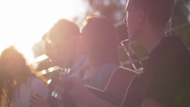 Jóvenes amigos caucásicos divirtiéndose y cantando canciones con una guitarra en el parque de verano al atardecer — Vídeo de stock