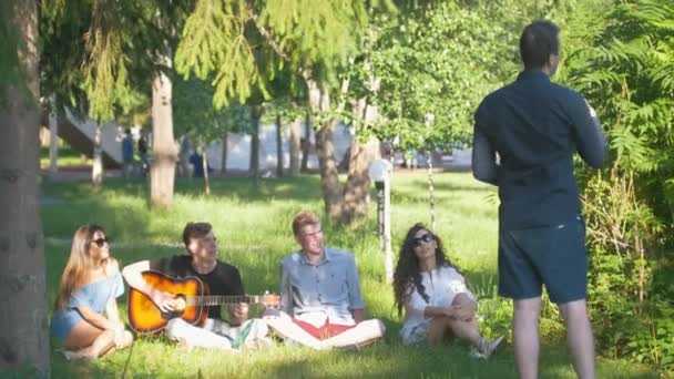 Jonge man toont pantomime voor vrienden zitten op het gras in de zomer park — Stockvideo