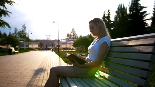 Aantrekkelijke jonge vrouw lezen van een boek, zittend op het bankje in het park bij zonsondergang — Stockvideo