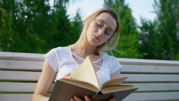 Muchacha atractiva leyendo un libro en el banco y quita el pelo de su cara — Vídeo de stock