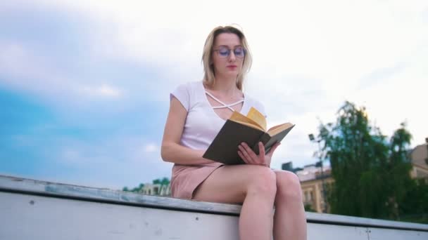 Attraktiv ung kvinna läser en bok utomhus, blå himmel i bakgrunden — Stockvideo