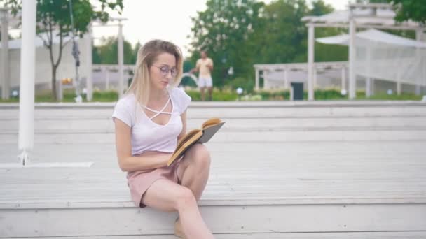 Junge Frau sitzt auf der Treppe und liest im Stadtpark ein Buch — Stockvideo