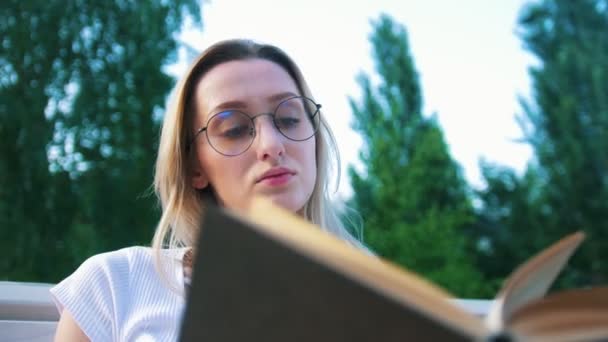 Πορτρέτο του ελκυστικό κορίτσι σε γυαλιά που κάθονται στον πάγκο διαβάζοντας ένα βιβλίο σε εξωτερικούς χώρους — Αρχείο Βίντεο