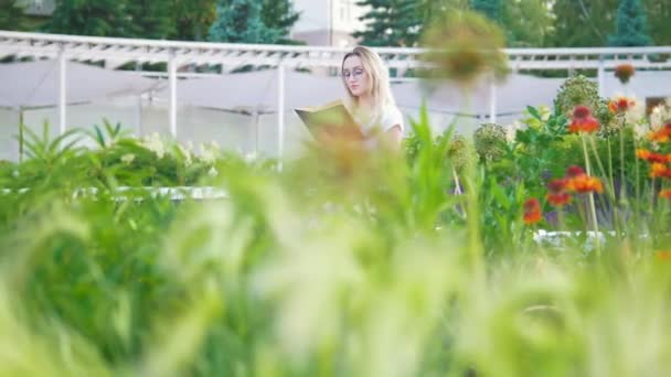 Νεαρή γυναίκα στον κήπο με τα λουλούδια που κάθονται στον πάγκο διαβάζοντας ένα βιβλίο — Αρχείο Βίντεο