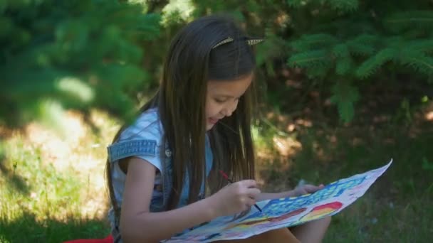 Menina bonito desenha imagens brilhantes com guache sentado na grama no parque de verão — Vídeo de Stock