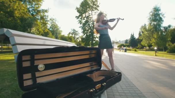 Estuche para violín tumbado en el banco frente a una hembra que toca un violín al atardecer — Vídeos de Stock