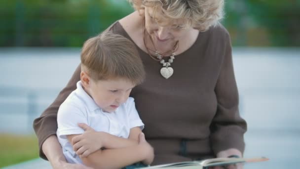 Boy and grandmother read book in the park on the bench — Stock Video