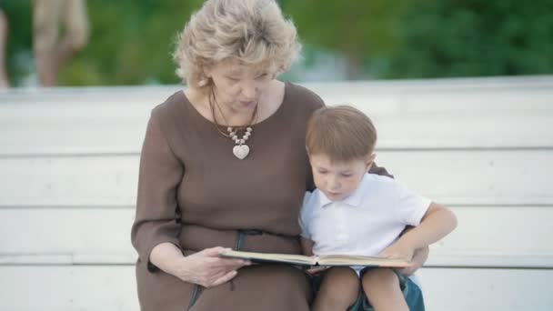 A boy with his grandmother reading and sitting on the white steps in the park — Stock Video