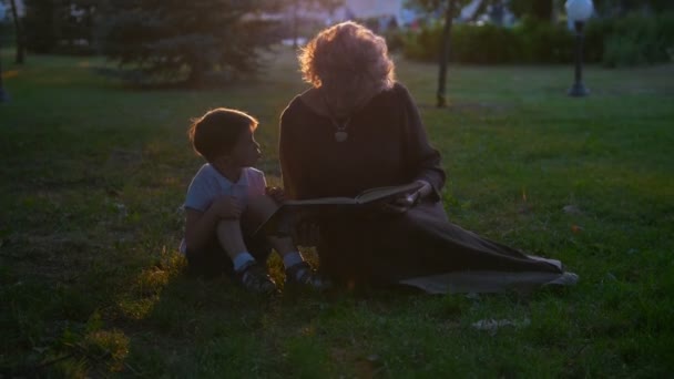 Grandmother reads a book to her grandson, sitting on the grass in the park at sunset — Stock Video