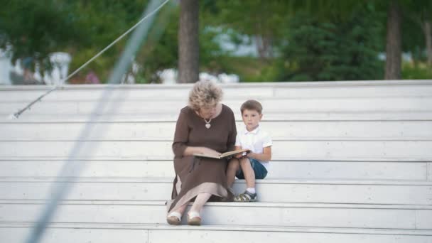 Abuela leyendo un libro a su nieto sentado en las escaleras en el parque — Vídeos de Stock