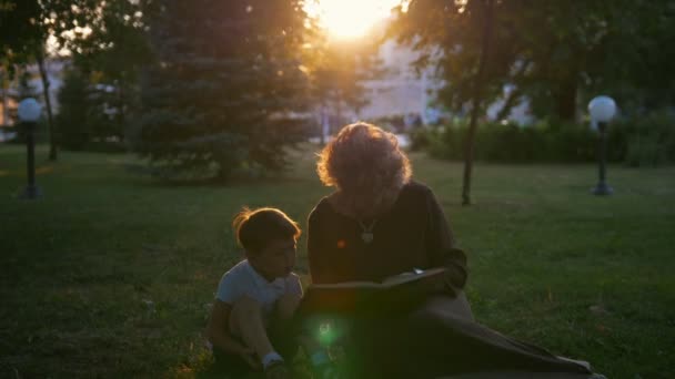Бабуся читає книгу онуку, що сидить на траві на заході сонця — стокове відео