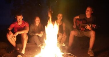 Kamp ateşi ve şarkıları söyleyerek bir gitarla oturan mutlu genç arkadaşlar