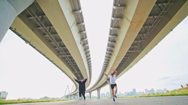 Два молодых человека делают акробатические трюки под мостом — стоковое видео