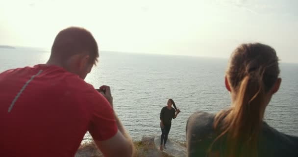 Rear view of young man shooting a beautiful woman stands on the edge of a cliff at riverside — Stock video