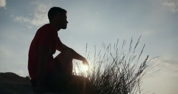 Silhouette di giovane seduto sul bordo di una scogliera e guardando il tramonto — Video Stock