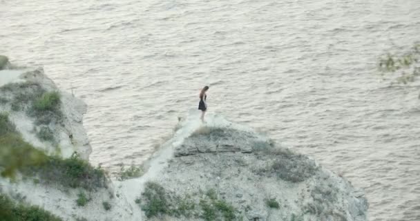 Jovem mulher de vestido preto dançando no penhasco junto ao mar — Vídeo de Stock