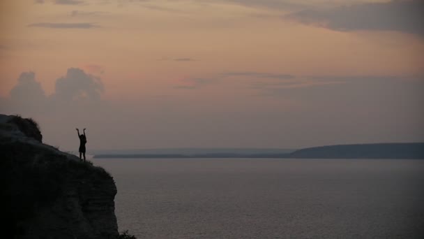 Silhouet van de vrouw met lange haren in zwarte jurk dansen op de rand van de klif bij zonsondergang — Stockvideo