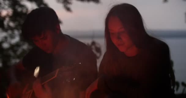 Jovem casal amoroso sentado ao lado da fogueira e tocando guitarra na floresta — Vídeo de Stock