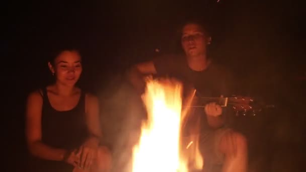 Jovens amigos cantando músicas com guitarra sentados ao lado da fogueira na noite escura — Vídeo de Stock