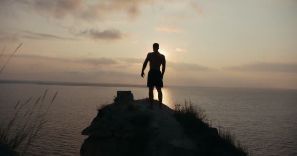 Silueta masculina haciendo sentadillas en la gran piedra junto al mar al amanecer — Vídeos de Stock