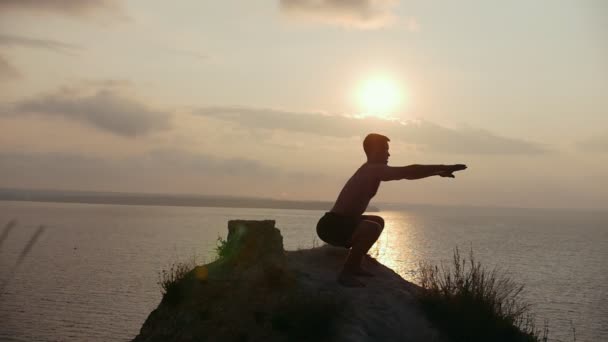 Jovem silhueta fazendo agachamentos no penhasco ao nascer do sol — Vídeo de Stock