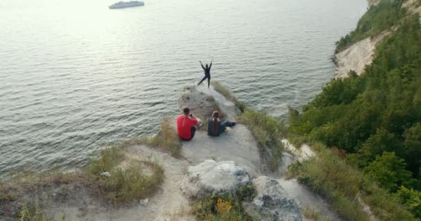 Jóvenes amigos sentados en la montaña mirando a la hermosa mujer bailando en el acantilado junto al mar — Vídeos de Stock