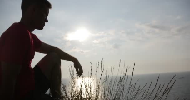 Joven sentado en el acantilado y mirando el atardecer — Vídeos de Stock