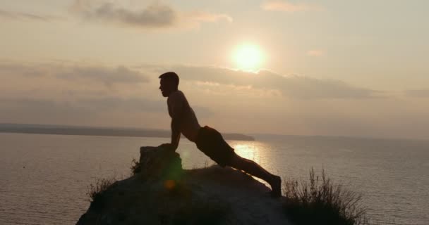 Hombre musculoso haciendo ejercicios matutinos en el acantilado junto al mar al amanecer — Vídeos de Stock