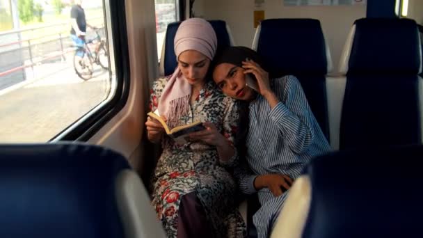 Zwei muslimische Frauen mit dem Zug unterwegs, eine liest, eine andere telefoniert — Stockvideo