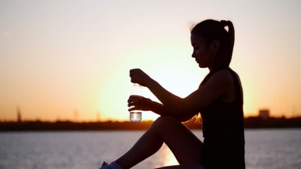 Deportiva joven y saludable bebiendo agua fría de la botella de plástico después de hacer ejercicio al atardecer — Vídeos de Stock