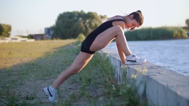 Jovem bela mulher treina suas pernas antes de correr na orla — Vídeo de Stock
