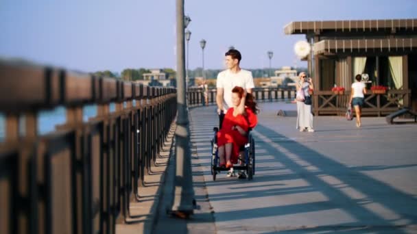 Guy Rolls A Disabled Una chica sonriente con el pelo rojo en una silla de ruedas en el paseo marítimo — Vídeo de stock