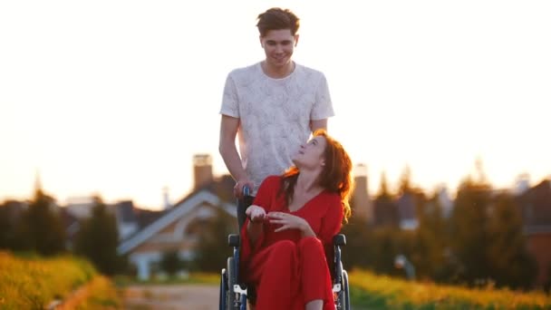 Улыбающийся молодой человек катит рыжеволосую девушку на инвалидном кресле, которая ему что-то говорит — стоковое видео