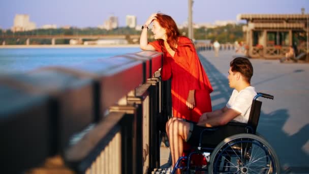 Chico en una silla de ruedas y chica con el pelo rojo se comunican en el paseo marítimo en la noche de verano — Vídeos de Stock