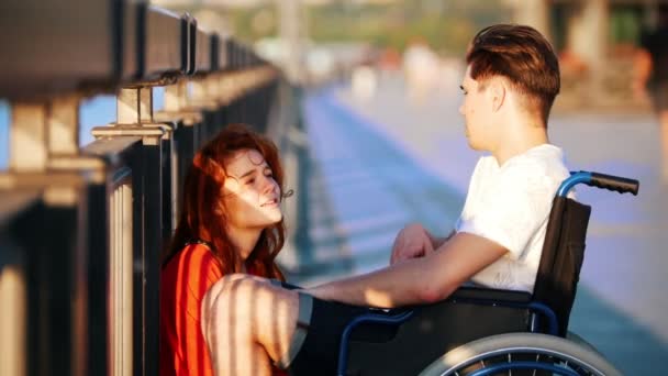 Guy On The Wheelchair Says Something To Red-Haired Girl And She Iistens Carefully, On Promenade In Summer Evening — Stock Video