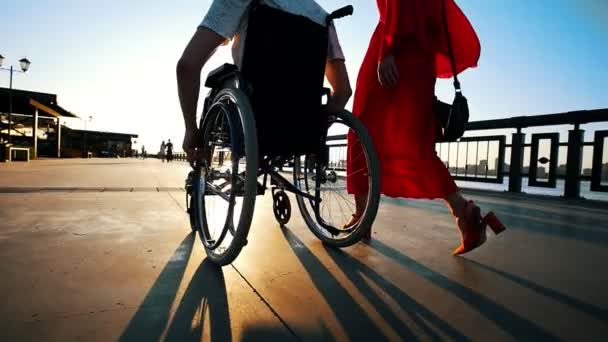 Cara com pernas doentes na cadeira de rodas andando com namorada no beira-mar na noite de verão — Vídeo de Stock