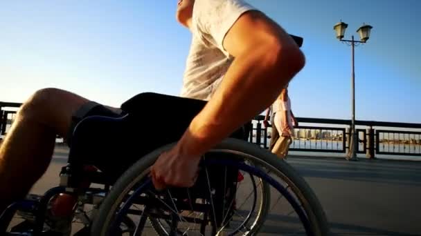 Guy monte en fauteuil roulant et une belle fille marche avec lui sur le front de mer au coucher du soleil — Video