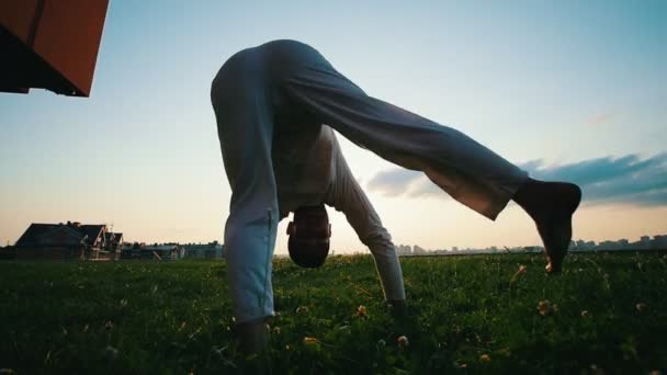 Athletic man is doing capoeira on the grass, summer evening at sunset — Stock Video