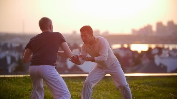 Sportliche Männer trainieren die Fertigkeiten des brasilianischen Tanzcapoeira vor dem Hintergrund der Stadt und eines wunderschönen Sonnenuntergangs — Stockvideo