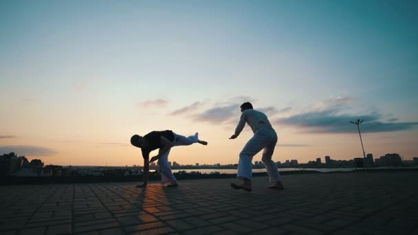 Män med en sportig fysik Dans capoeira utomhus, på trottoaren, mot den vackra strandpromenaden och sommar solnedgång — Stockvideo