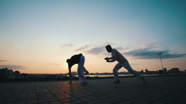Homens com um físico esportivo estão envolvidos na arte marcial brasileira da capoeira ao ar livre, no asfalto, no fundo da beleza do passeio e no pôr-do-sol de verão. — Vídeo de Stock